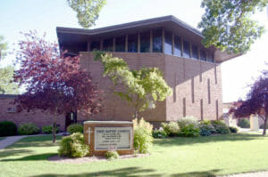 Exterior of brown-bricked church with three-story worship hall with windows on top story and First Baptist Church sign in the foreground.