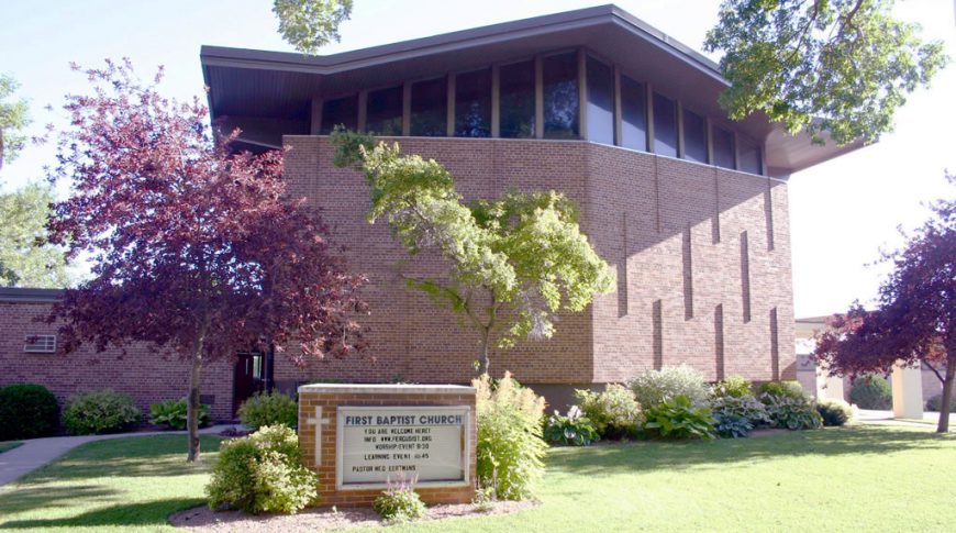 Exterior of brown-bricked church with three-story worship hall with windows on top story and First Baptist Church sign in the foreground.