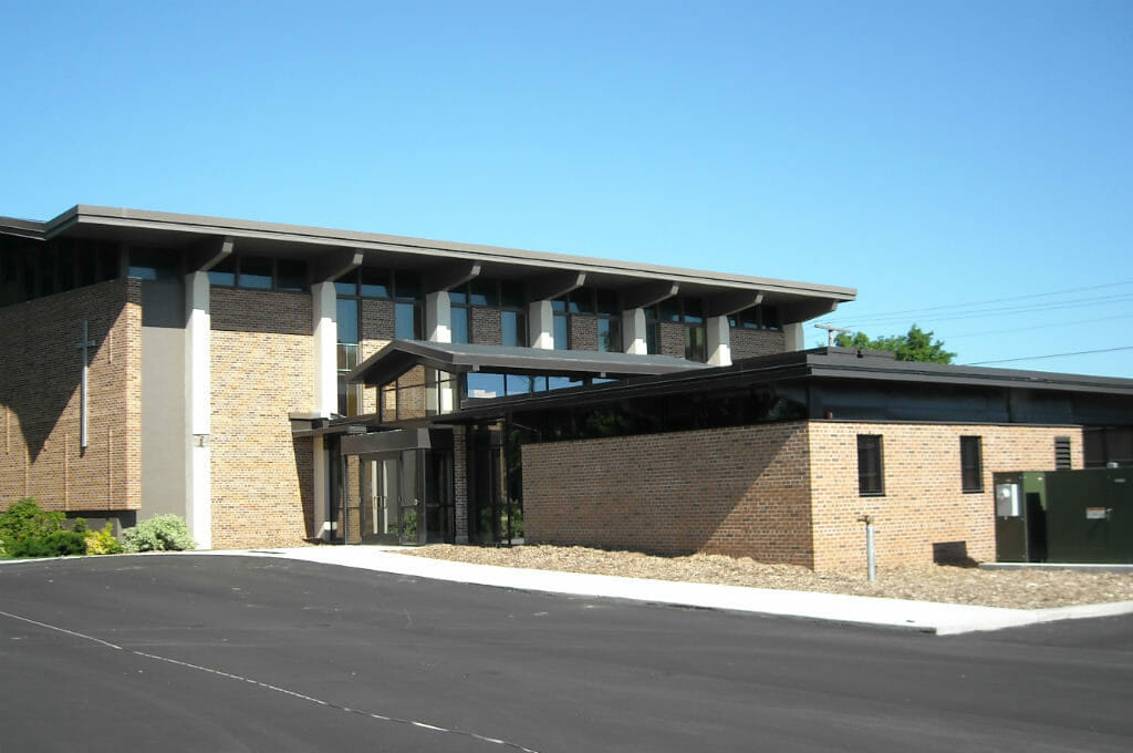 Bi-level brown brick building with glass entrance on lower half and asphalt parking lot in front.