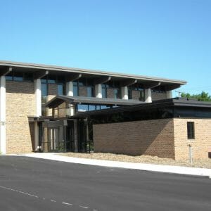 Bi-level brown brick building with glass entrance on lower half and asphalt parking lot in front.