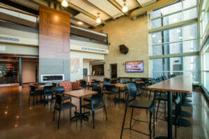 Two-story common room with short and tall tables and chairs with two-story modern fireplace and windows on other side.