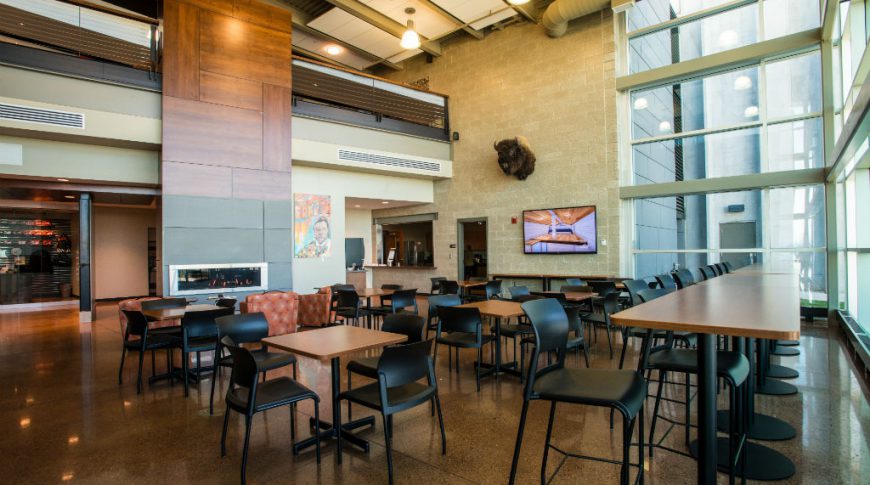 Bobcat Acceleration Center-P-Int1-1024x680-Commercial Construction Two-story common room with short and tall tables and chairs with two-story modern fireplace and windows on other side.