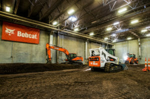 Large interior space with dirt floor and concrete walls and overhead metal roof and exposed beams with Bobcat equipment and Bobcat banner on back wall.