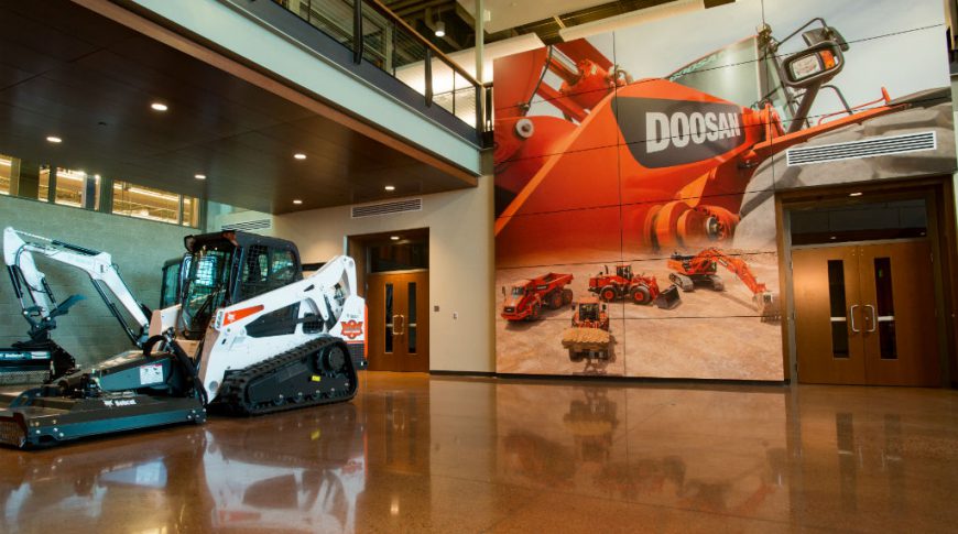 Bobcat Acceleration Center-P-Int5-1024x680-Commercial Construction Concrete speckled flooring with bobcat machinery to back left and floor-to-ceiling paneled accent wall with picture of Doosan equipment on back wall with second-floor mezzanine overlooking.