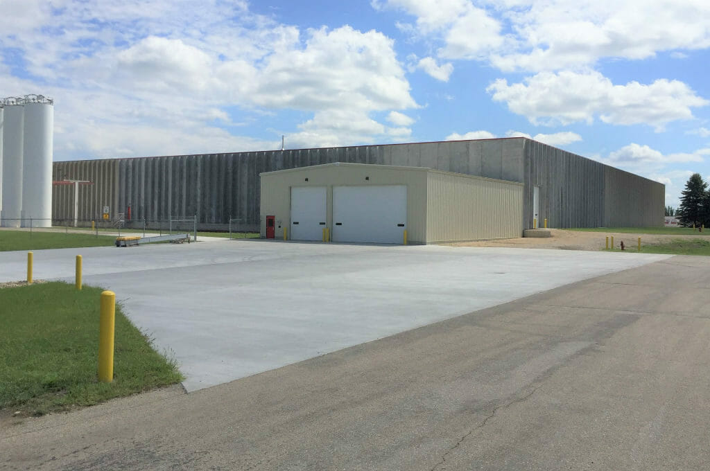 Large concrete rectangular building with smaller rectangular metal beige building attached in the front with a couple truck bays and oil tanks to the left.