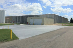 Large concrete rectangular building with smaller rectangular metal beige building attached in the front with a couple truck bays and oil tanks to the left.