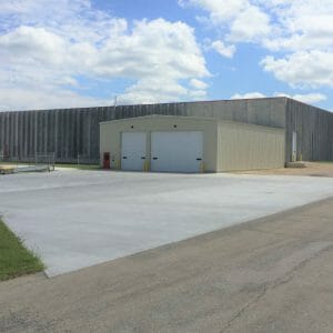 Large concrete rectangular building with smaller rectangular metal beige building attached in the front with a couple truck bays and oil tanks to the left.