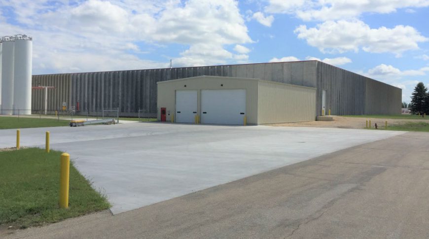 ComDel Innovation 1Exp-Ext2-edit2-Commercial Construction Large concrete rectangular building with smaller rectangular metal beige building attached in the front with a couple truck bays and oil tanks to the left.