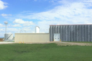 Exterior of concrete building with one garage bay, attached to beige metal building on left side with oil tanks in the background.