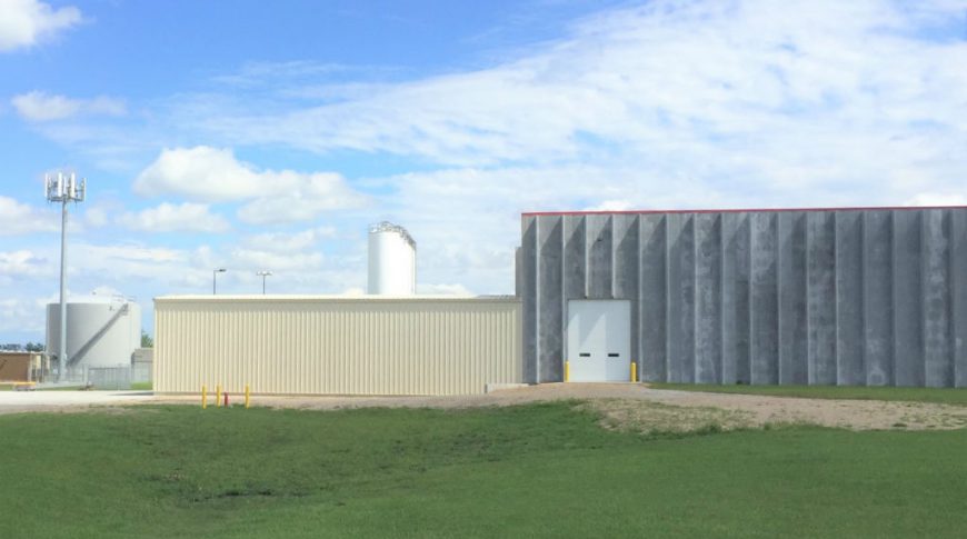 ComDel Innovation 1Exp-Ext3-edit2-Commercial Construction Exterior of concrete building with one garage bay, attached to beige metal building on left side with oil tanks in the background.