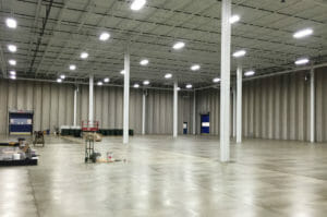 Open warehouse with concrete floors and exposed white ceilings and trusses with fluorescent lights overhead. Concrete walls and white steel beams throughout.