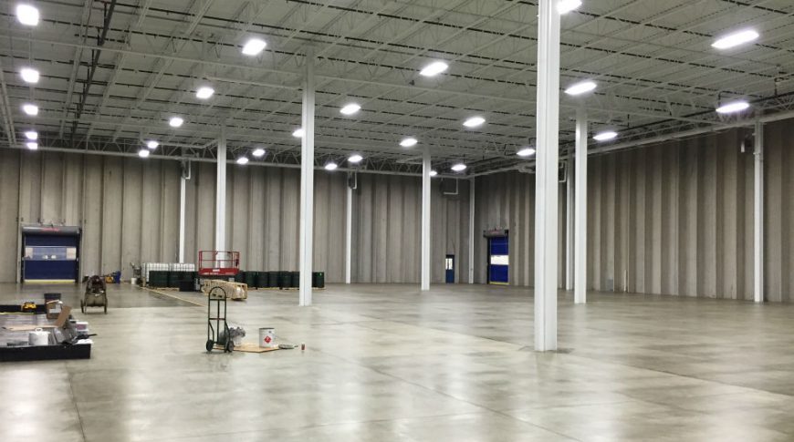 ComDel Innovation1Exp-Int4-edit-Commercial Construction Open warehouse with concrete floors and exposed white ceilings and trusses with fluorescent lights overhead. Concrete walls and white steel beams throughout.