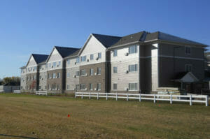 Backside view of three-story beige and brown-bricked residence building with white accent fence to the foreground.