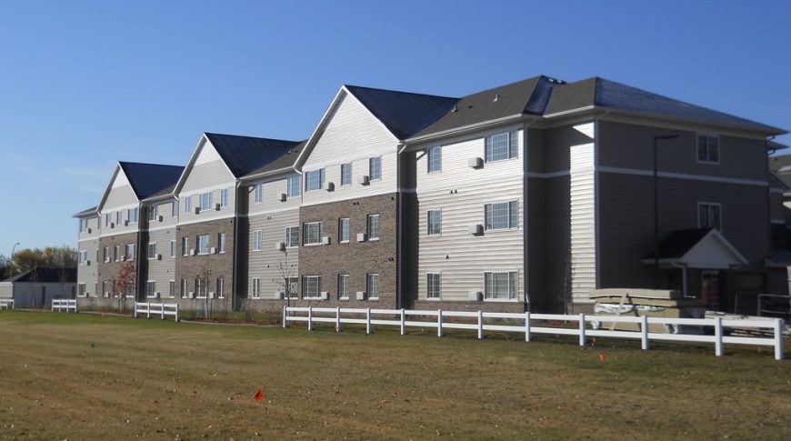 Backside view of three-story beige and brown-bricked residence building with white accent fence to the foreground.