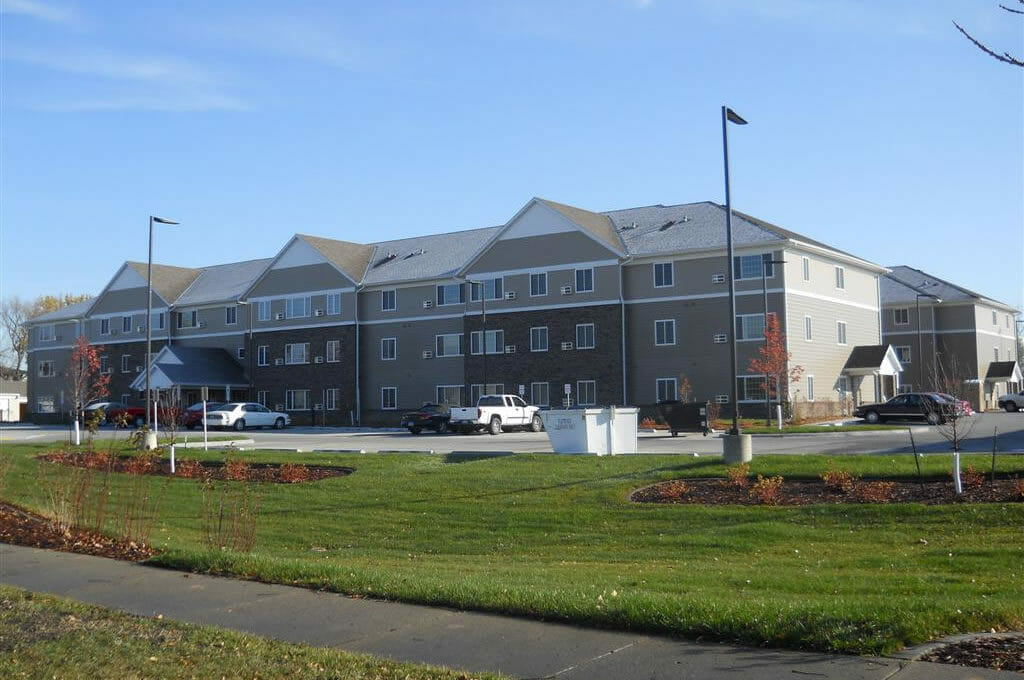 Exterior road view of three-story beige and brown bricked senior living community with parking lot in foreground.