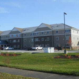 Exterior road view of three-story beige and brown bricked senior living community with parking lot in foreground.