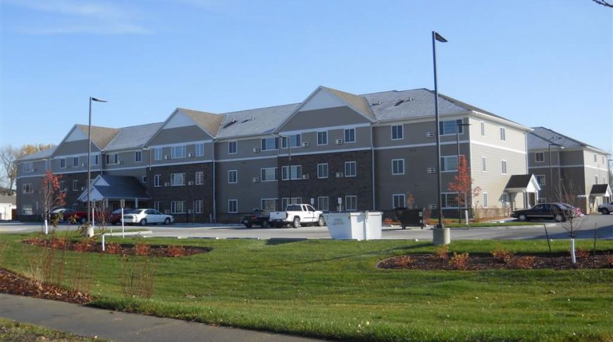 Exterior road view of three-story beige and brown bricked senior living community with parking lot in foreground.