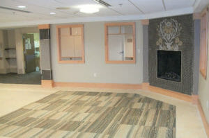 Room with light gray walls, tiled fireplace surround in back right corner, light wooden trim around baseboards and windows and light beige and blue carpet tiles inset around ceramic floor tiles.