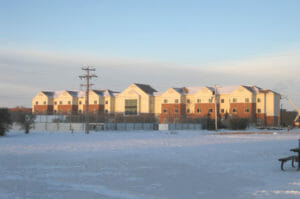 Far away view of entire F08 dormitory building on the Minot Air Force Base on a winter day. Snow in foreground with cream and red-bricked three-story dormitory building in background.