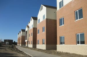 Exterior of three-story red-bricked FY08 dormitory building with light cream siding on the top floor. Two windows spaced on each level with unfinished sidewalk and landscaping beneath.