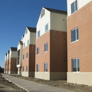 Exterior of three-story red-bricked FY08 dormitory building with light cream siding on the top floor. Two windows spaced on each level with unfinished sidewalk and landscaping beneath.