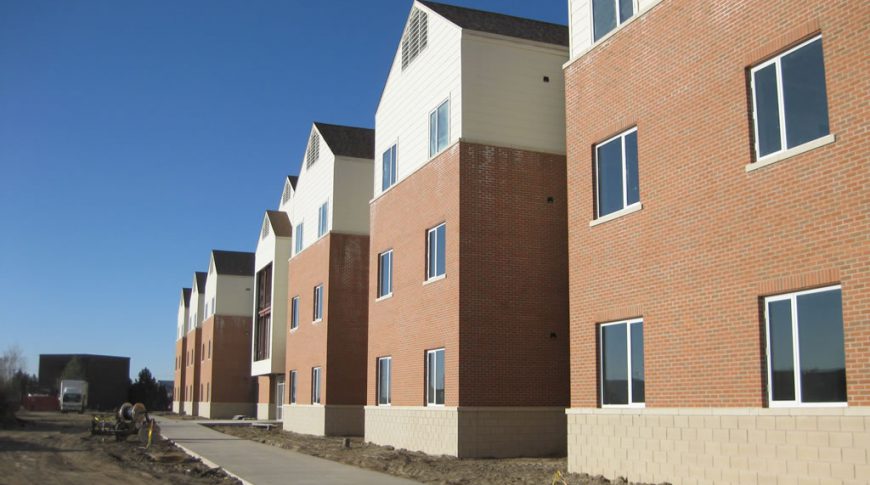Exterior of three-story red-bricked FY08 dormitory building with light cream siding on the top floor. Two windows spaced on each level with unfinished sidewalk and landscaping beneath.
