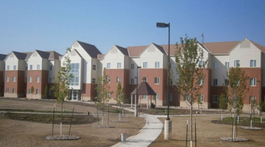 View from sidewalk of three story FY08 Dormitory housing buildings in beige and red siding with windows throughout overlooking park with gazebo and sidewalks.