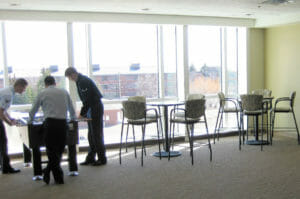 Three men playing foosball in back left corner with two high-top cocktail tables and chair to the right of them with a wall of windows behind at fy08 dormitory.