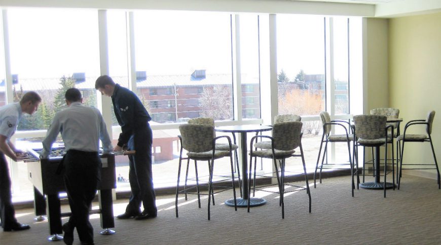 Three men playing foosball in back left corner with two high-top cocktail tables and chair to the right of them with a wall of windows behind.