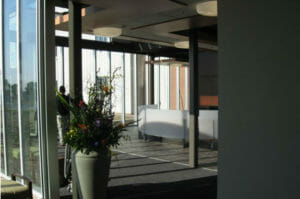 Entrance to building with large concrete flower pot with brightly colored flowers inside viewing inside of room with floor-to-ceiling windows on two sides and large semi-circle custom couch in center.