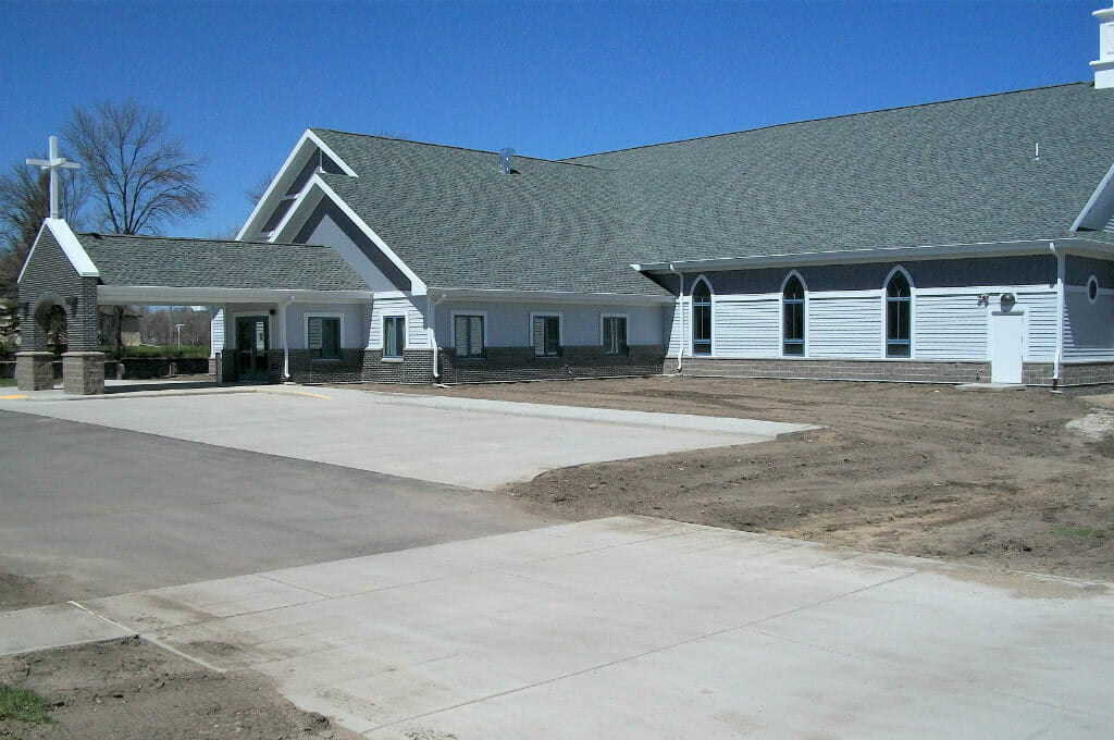 Light gray and dark gray church building with charcoal roof shingles and covered carport with cross on top.