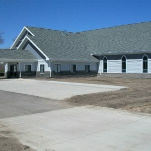 Light gray and dark gray church building with charcoal roof shingles and covered carport with cross on top.