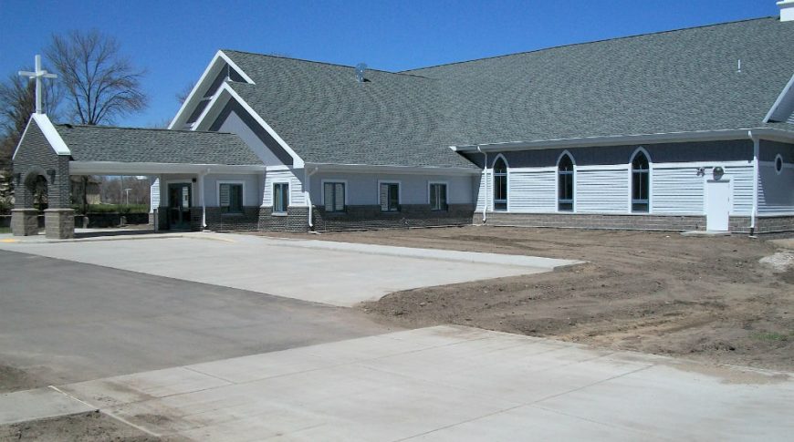 Light gray and dark gray church building with charcoal roof shingles and covered carport with cross on top.