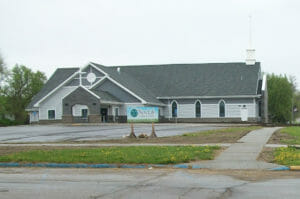 Road view of light and dark gray church with white steeple cross and cross over carport.