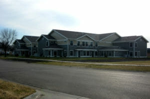 Across the street view of two-story assisted living building with beige and gray siding with brown shingled roof. Unfinished landscaping in front.