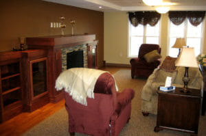 Assisted living living room area with beige couch and two burgundy armchairs on either side facing fireplace and wooden mantle with built-ins on either side.
