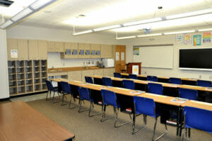 Large beige classroom with cabinetry on back left wall with whiteboard and smart board on back right wall. Desks and tables seated classroom style in foreground.