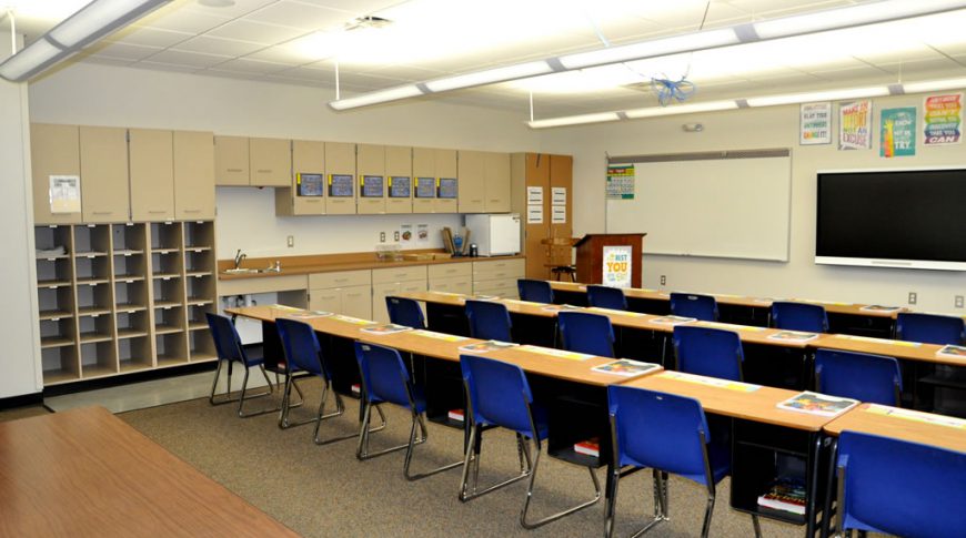 Stanley Elementary School-Int3-Education Construction Large beige classroom with cabinetry on back left wall with whiteboard and smart board on back right wall. Desks and tables seated classroom style in foreground.