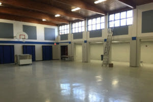 Interior of cafeteria that doubles as a gymnasium with laminate floors and two-story high ceiling with wooden sheeting and beams and windows on second story.