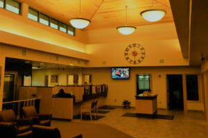 Bank foyer with seating chairs to the bottom left and high wood paneled ceiling with windows overhead and teller stations to the back left.