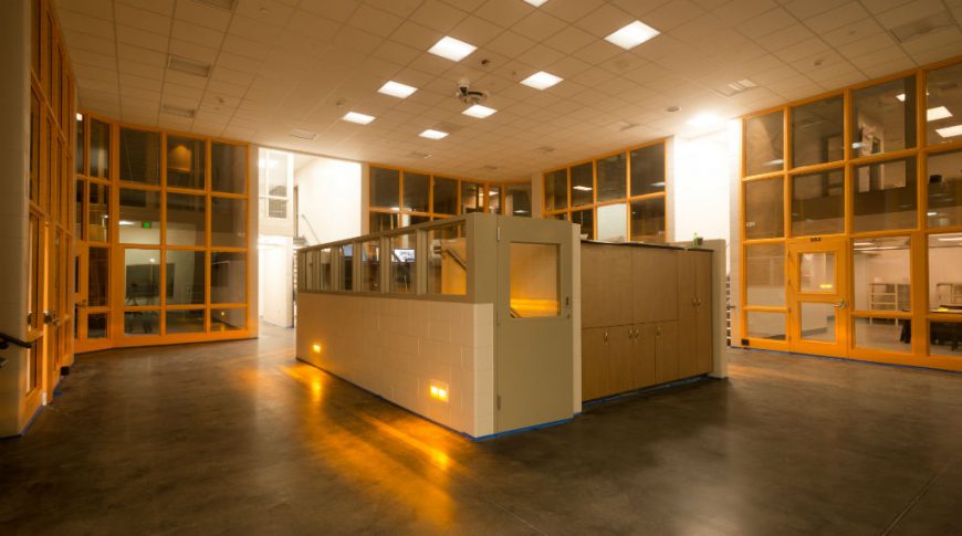 Burleigh Morton Detention Center-P-Int3-1024x680-Government Construction Two-story room with concrete floors, center cinder block and light cabinetry station all surrounded by wall of windows on all sides.