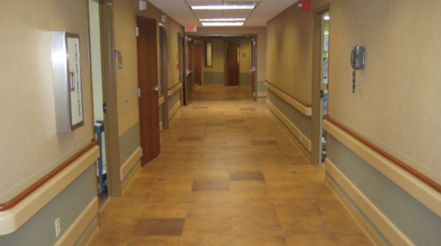 Cavalier County Memorial Hospital-Int9-1024x680-Healthcare Construction Hospital hallway with beige tiled floor underneath and blue and beige colored walls with chair ledge on each side and doorways to rooms on each side.