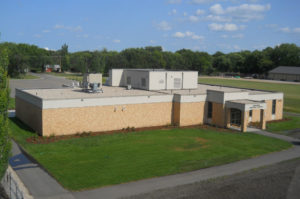 Aerial view of light beige brick building with concrete awning entrance surrounded by lush green grass and trees with bright blue sky overhead.