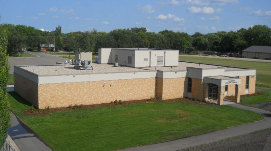 Aerial view of light beige brick building with concrete awning entrance surrounded by lush green grass and trees with bright blue sky overhead.