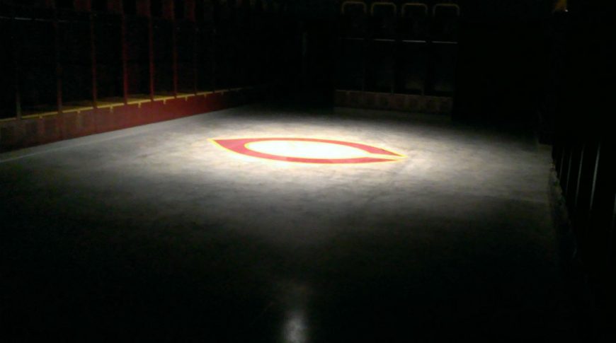 Dimly lit Concordia locker room with spotlight on concrete floor with the C logo and glimpses of maroon and gold lockers around the edges.