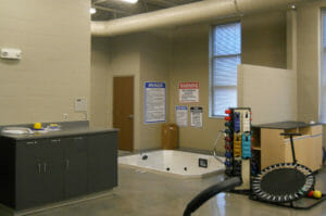 Therapy room with in-ground jetted hot tub with countertop station to left wall and small ankle weights, dumbbells and trampoline station to right.