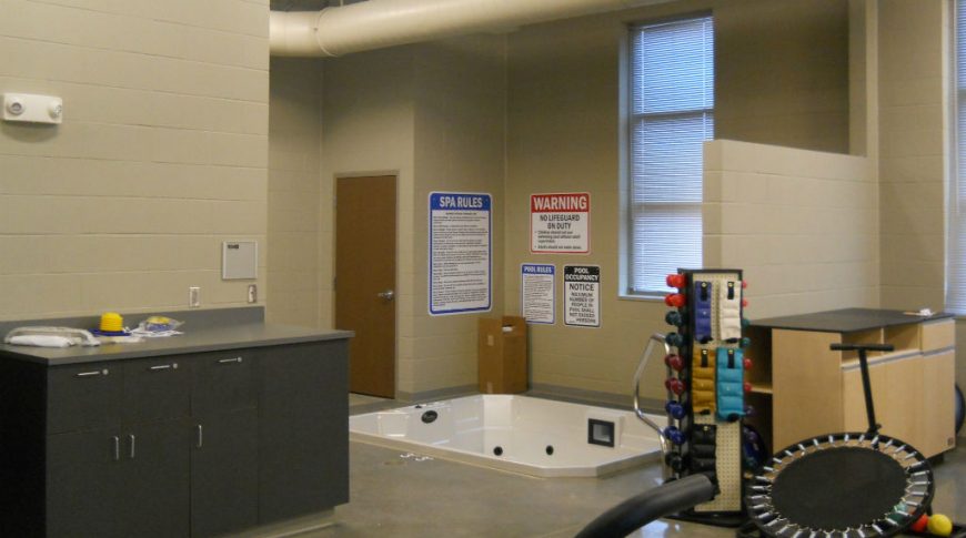 Therapy room with in-ground jetted hot tub with countertop station to left wall and small ankle weights, dumbbells and trampoline station to right.