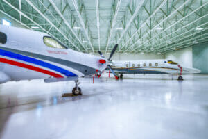 Interior of hangar building with light white floors and white metal ceiling overhead with fluorescent lights and private jets throughout.