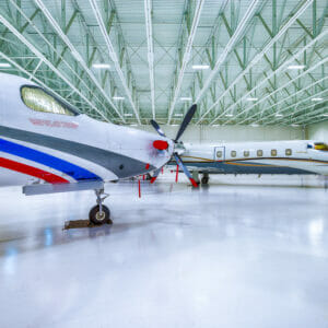 Interior of hangar building with light white floors and white metal ceiling overhead with fluorescent lights and private jets throughout.