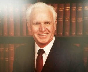 Head and shoulders portrait of owner Lynn Comstock who is an older white male with white hair wearing black suite, white-collared shirt and dark maroon and polka dot tie with library book backdrop